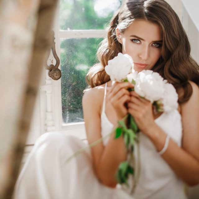 Adorable young bride holds a bouuqet of peonies siting on the wi