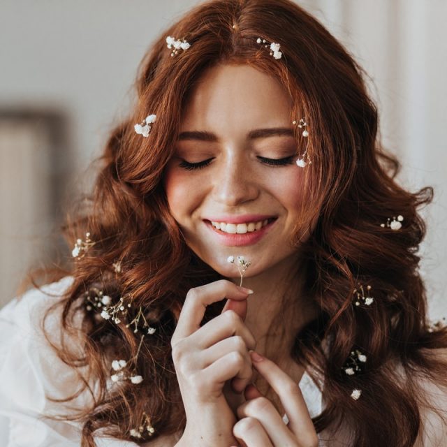 Lady with flowers in red curls with smile posing. Indoor portrait of girl with small plant in her hands.